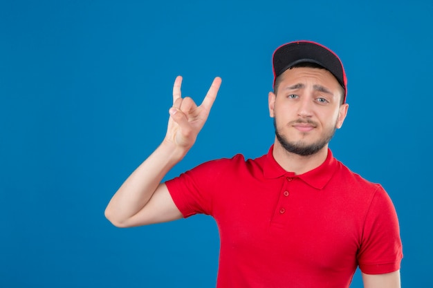 Junger Lieferbote, der rotes Poloshirt und Kappe trägt Rocksymbol, das zuversichtlich über lokalisiertem blauem Hintergrund schaut