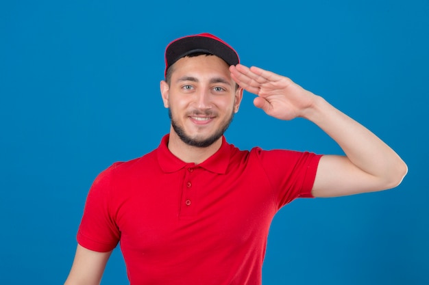 Junger Lieferbote, der rotes Poloshirt und Mütze salutierend schaut, die über lokalem blauem Hintergrund sicher schauen