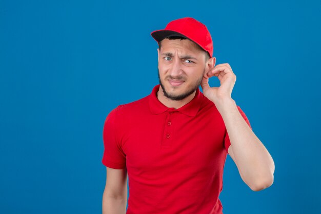 Junger Lieferbote, der rotes Poloshirt und Mütze trägt, die Kamera betrachten, die sein Ohr berührt, hört nicht jemandem zu, der über isoliertem blauem Hintergrund liegt