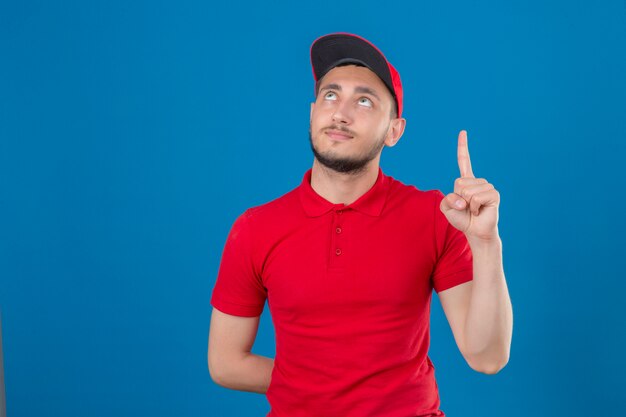 Junger Lieferbote, der rotes Poloshirt und Mütze trägt, die zuversichtlich mit dem Finger zeigt, der über lokalisiertem blauem Hintergrund zeigt