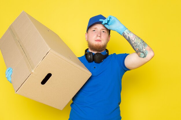 junger Lieferbote im blauen Jeans-Rucksack der blauen Polo-blauen Kappe, der eine Schachtel auf Gelb hält