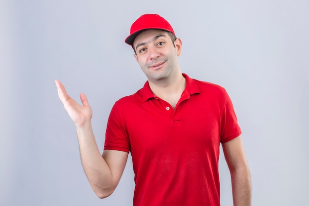 Junger Lieferbote im roten Poloshirt und in der Kappe, die etwas mit Hand über lokalisierten weißen Hintergrund präsentieren