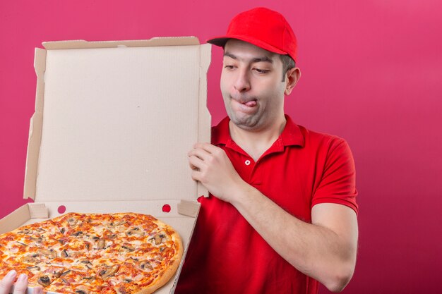 Junger Lieferbote im roten Poloshirt und in der Kappe, die mit der Schachtel der frischen Pizza stehen, die es mit einem hungrigen lustvollen Gesicht über lokalisiertem rosa Hintergrund betrachtet