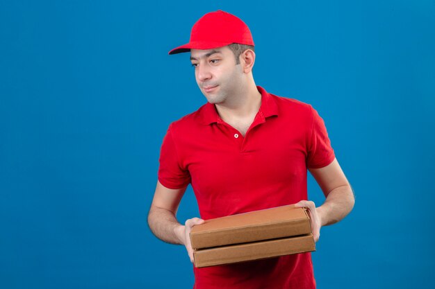 Junger Lieferbote im roten Poloshirt und in der Kappe, die Pizzaschachteln halten, die zur Seite mit ernstem Gesicht über isolierter blauer Wand schauen