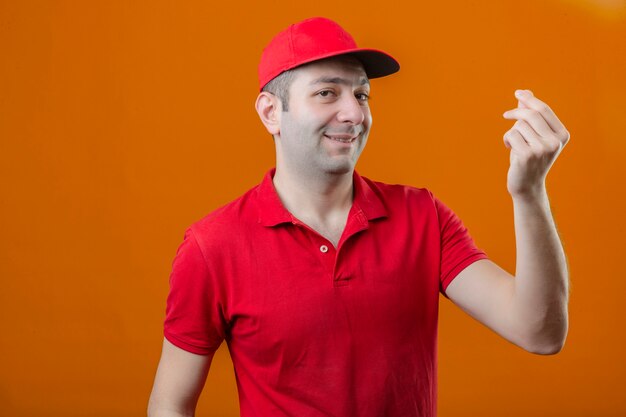 Junger Lieferbote im roten Poloshirt und in der Kappe mit dem Lächeln auf Gesicht, das Geldgestik mit Hand über isolierte orange Wand macht