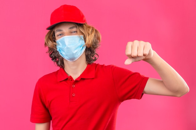 Junger Lieferbote in der roten Uniform, die medizinische Maske trägt, die gestikulierende Faustbeule zwinkert, als ob Gruß genehmigt oder als Zeichen des Respekts über isoliertem rosa Hintergrund