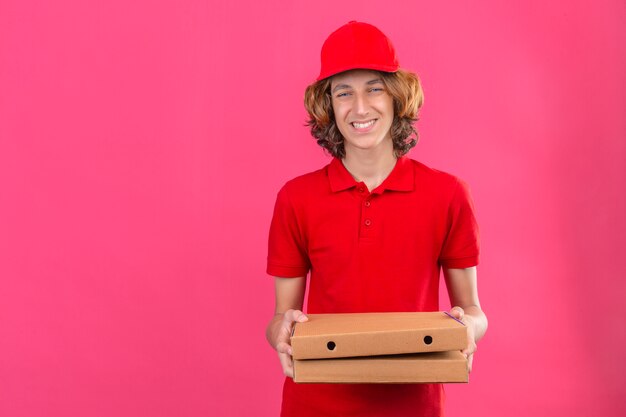 Junger Lieferbote in der roten Uniform, die Pizzakästen hält, die fröhlich mit glücklichem Gesicht über lokalisiertem rosa Hintergrund lächeln