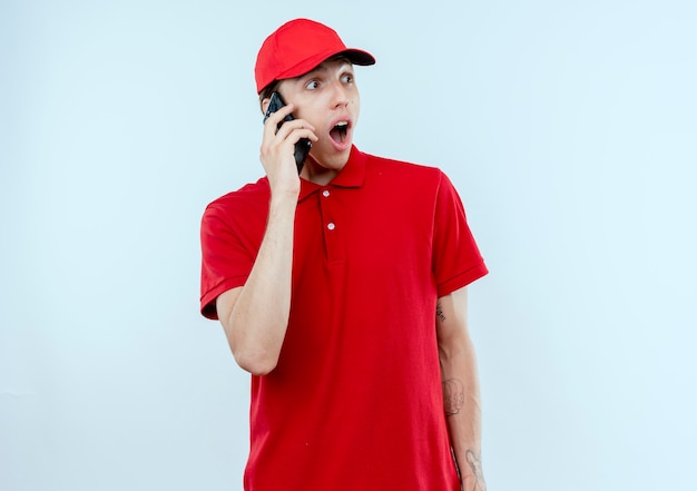Junger Lieferbote in der roten Uniform und in der Kappe, die auf dem schockierten Ausdruck des Mobiltelefons über weißer Wand stehen