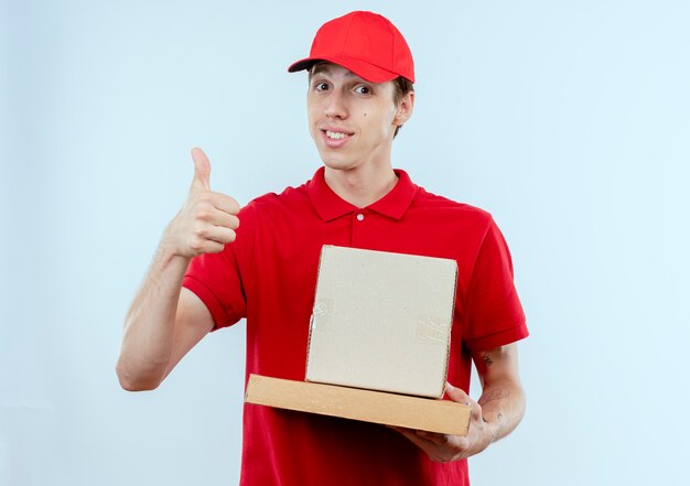 Junger Lieferbote in der roten Uniform und in der Kappe, die Boxpaket und in der Pizzaschachtel hält, die nach vorne lächelnd zeigt Daumen hoch stehend über weißer Wand