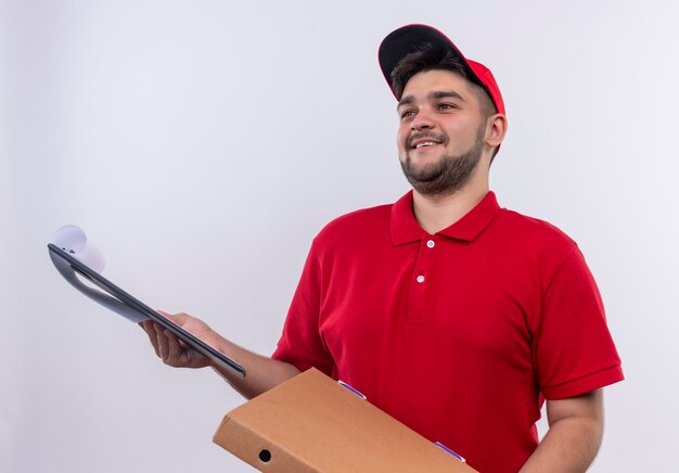 Junger Lieferbote in der roten Uniform und in der Kappe, die Boxpaket und Zwischenablage mit leeren Seiten hält, die um Unterschrift bitten