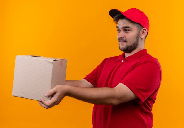 Junger Lieferbote in der roten Uniform und in der Kappe, die Boxpaket zu einem Kunden geben, der freundlich lächelt