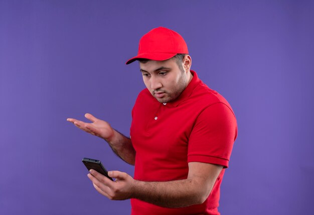 Junger Lieferbote in der roten Uniform und in der Kappe, die den Bildschirm seines Smartphones verwirrt und enttäuscht betrachten, über lila stehend