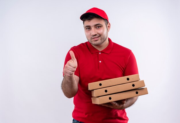 Junger Lieferbote in der roten Uniform und in der Kappe, die die Pizzaschachteln lächelnd freudig zeigend zeigt Daumen hoch stehend über weißer Wand