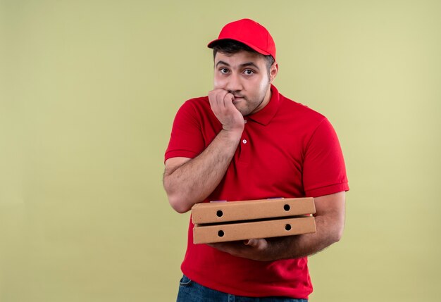 Junger Lieferbote in der roten Uniform und in der Kappe, die gestresste und nervöse beißende Pizzakästen hält, die über grüner Wand stehen