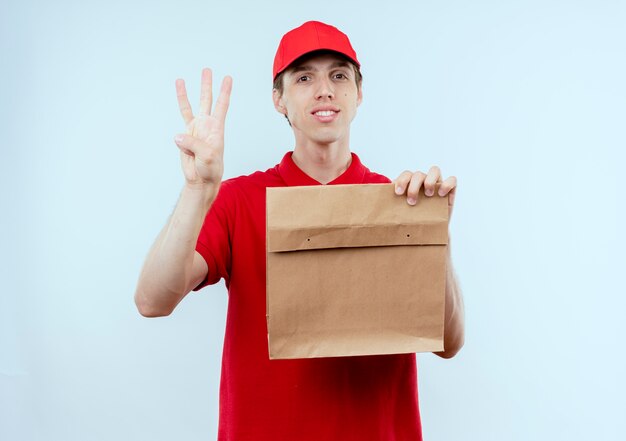 Junger Lieferbote in der roten Uniform und in der Kappe, die Papierpaket hält, das Nummer drei zeigt, das zuversichtlich steht, über weißer Wand zu stehen