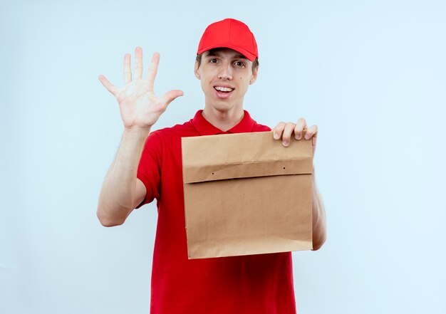 Junger Lieferbote in der roten Uniform und in der Kappe, die Papierpaket halten, das das lächelnde selbstbewusste Stehen Nummer fünf über weißer Wand zeigt