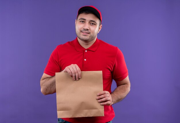 Junger Lieferbote in der roten Uniform und in der Kappe, die Papierpaket mit Lächeln auf Gesicht hält, das über lila Wand steht