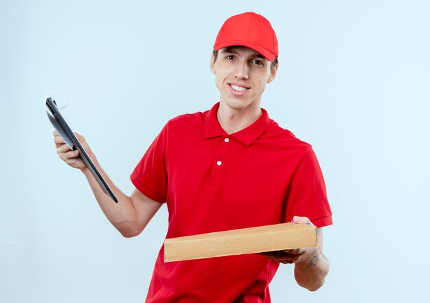 Junger Lieferbote in der roten Uniform und in der Kappe, die Pizzaschachtel und Zwischenablage hält, die zuversichtlich stehen über weißer Wand stehen