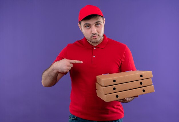 Junger Lieferbote in der roten Uniform und in der Kappe, die Pizzaschachteln halten, die mit dem Finger auf sie zeigen, die zuversichtlich stehen, über lila Wand stehen
