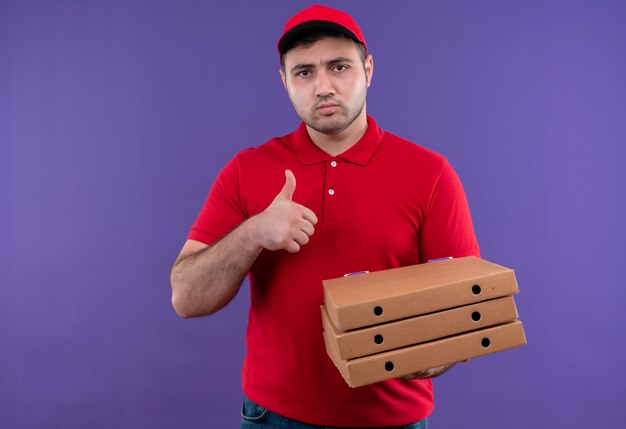 Junger Lieferbote in der roten Uniform und in der Kappe, die Pizzaschachteln mit stirnrunzelndem Gesicht hält, das Daumen oben zeigt über lila Wand