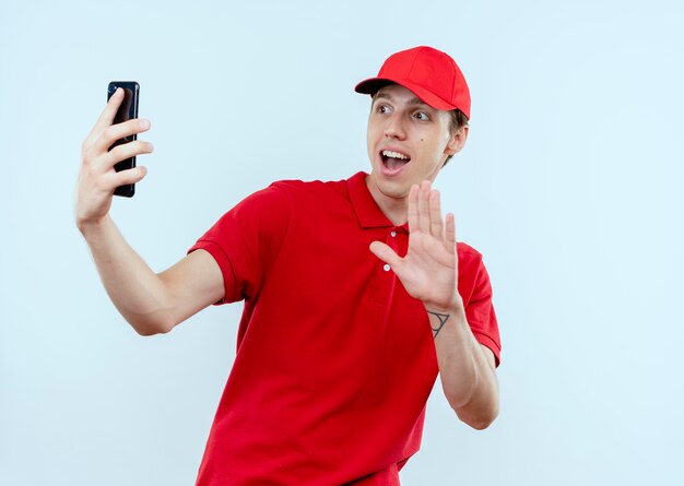 Junger Lieferbote in der roten Uniform und in der Kappe, die Smartphone hält, das das lächelnde Selfie winkend mit einer Hand hält, die über weißer Wand steht