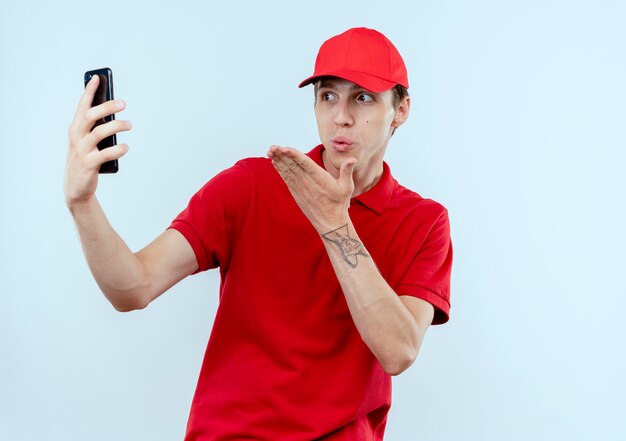 Junger Lieferbote in der roten Uniform und in der Kappe, die Smartphone halten, das lächelndes Selfie nimmt, das einen Kuss weht, der über weißer Wand steht