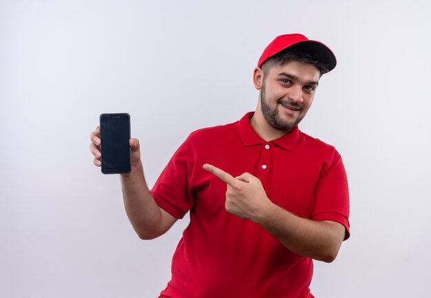 Junger Lieferbote in der roten Uniform und in der Kappe, die Smartphone zeigt, das mit dem Finger darauf zeigt, der fröhlich lächelt