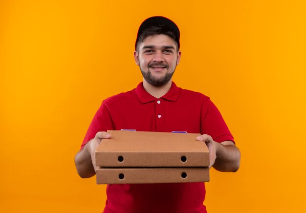 Junger Lieferbote in der roten Uniform und in der Kappe, die Stapel von Pizzaschachteln hält, die zuversichtlich lächeln