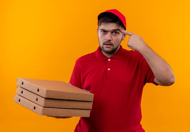 Junger Lieferbote in der roten Uniform und in der Kappe, die Stapel von Pizzaschachteln halten, die seine Schläfe mit ernstem Gesicht zeigen, das auf Aufgabe konzentriert ist