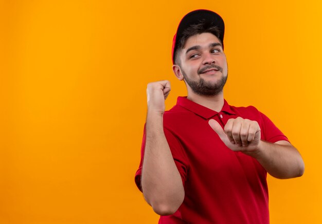 Junger Lieferbote in der roten Uniform und in der Kappe, die zurück lächelnd zuversichtlich zeigen