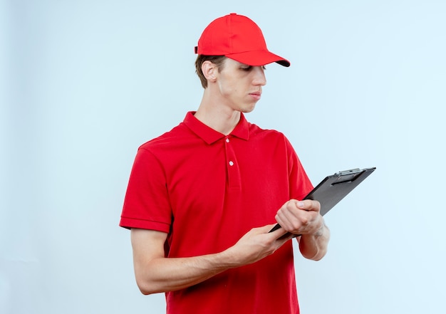 Junger Lieferbote in der roten Uniform und in der Kappe, die Zwischenablage hält, die es mit ernstem Gesicht betrachtet, das über weißer Wand steht