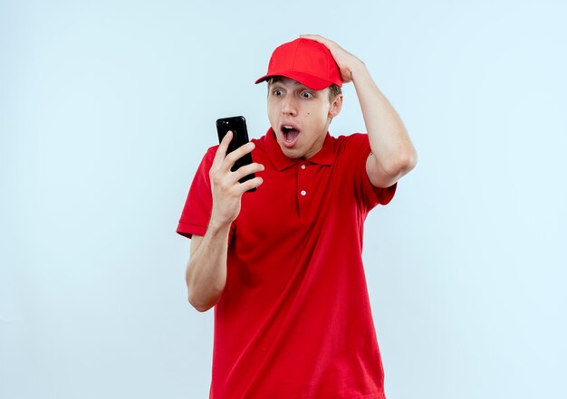Junger Lieferbote in der roten Uniform und in der Mütze, die den Bildschirm seines Handys betrachten, überrascht und verwirrt mit der Hand auf seinem Kopf, der über weißer Wand steht