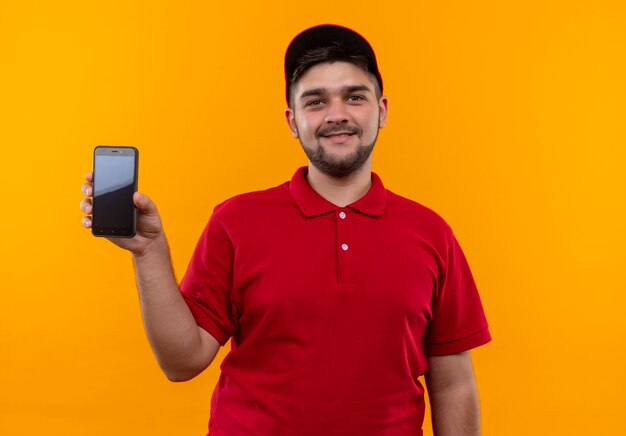 Junger Lieferbote in roter Uniform und Kappe, die Smartphone zeigt, das mit Lächeln auf Gesicht zuversichtlich schaut