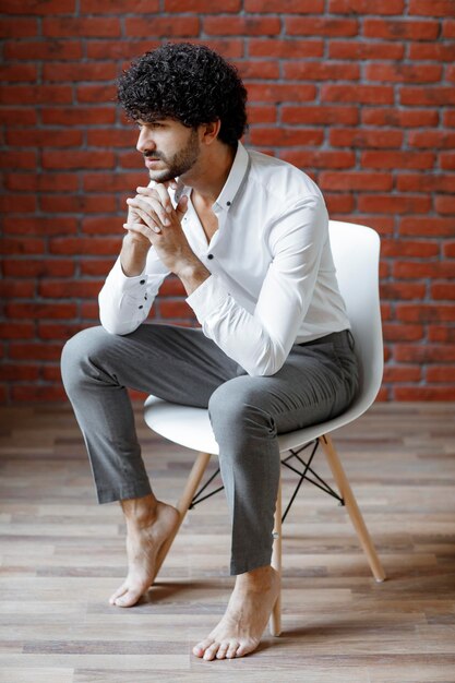 Junger, lockiger, gutaussehender Mann in weißem Hemd, der auf einem weißen Stuhl sitzt. Foto in hoher Qualität
