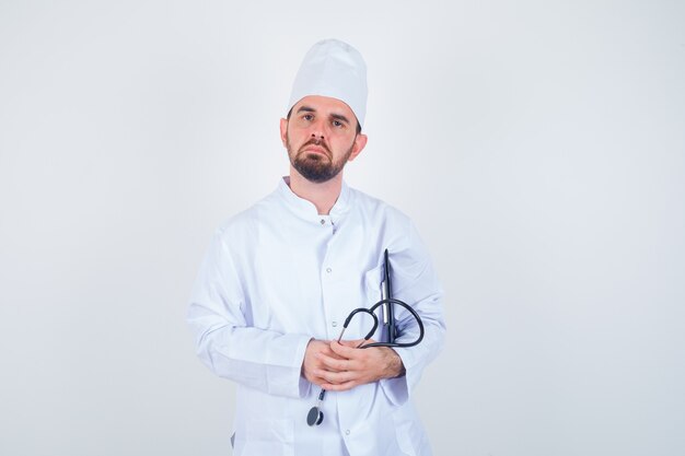 Junger männlicher Arzt, der Klemmbrett und Stethoskop in der weißen Uniform hält und vorsichtig schaut, Vorderansicht.