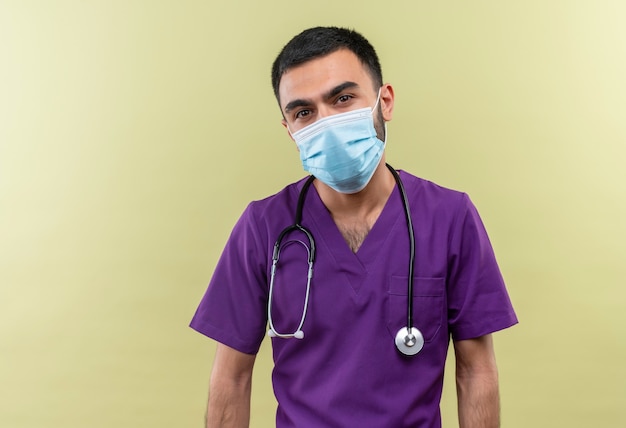 junger männlicher Arzt, der lila Chirurgenkleidung und medizinische Stethoskopmaske auf isolierter grüner Wand trägt