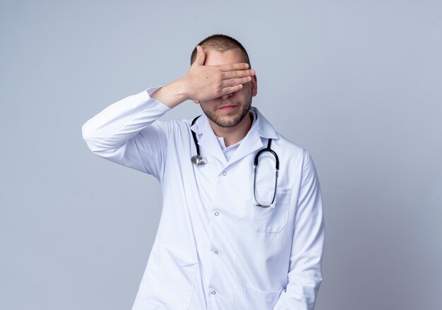 Junger männlicher Arzt, der medizinische Robe und Stethoskop um seinen Hals trägt, der Augen mit Hand lokalisiert auf weißem Hintergrund mit Kopienraum schließt