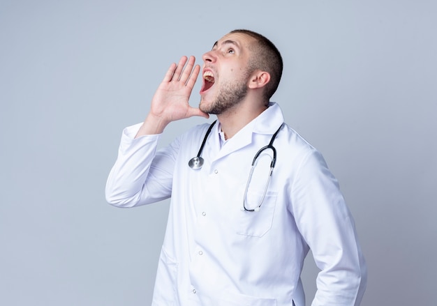 Junger männlicher Arzt, der medizinische Robe und Stethoskop um seinen Hals trägt, schaut auf und ruft laut zu jemandem mit Hand nahe Mund lokalisiert auf weißem Hintergrund mit Kopienraum