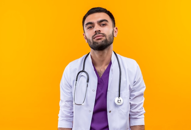 junger männlicher Arzt, der medizinisches Kleid des Stethoskops auf isolierter gelber Wand trägt