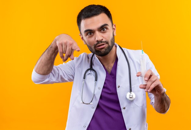 junger männlicher Arzt, der medizinisches Kleid des Stethoskops hält Spritze hält, die Sie Geste auf isolierter gelber Wand zeigt