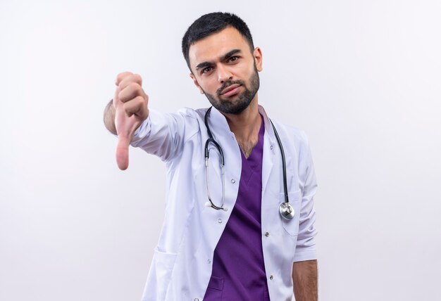 junger männlicher Arzt, der medizinisches Kleid des Stethoskops seinen Daumen unten auf isolierter weißer Wand trägt
