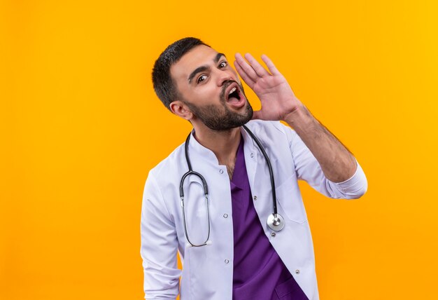 junger männlicher Arzt, der medizinisches Kleid des Stethoskops trägt, flüstert auf isolierter gelber Wand