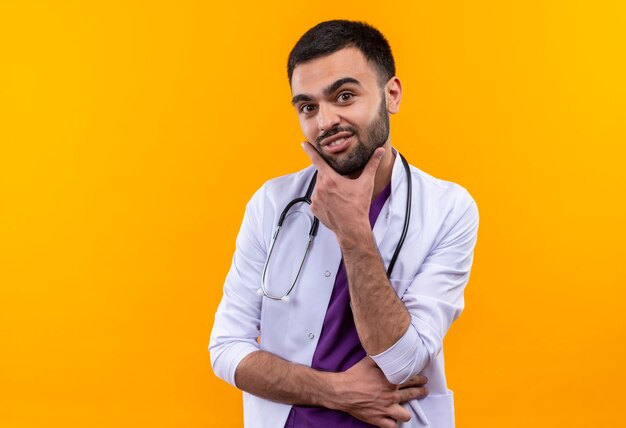 junger männlicher Arzt, der medizinisches Kleid des Stethoskops trägt, legte seine Hand auf Kinn auf isolierte gelbe Wand