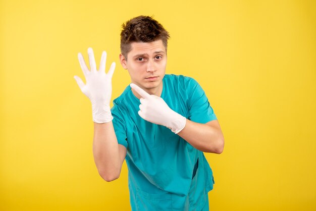 Junger männlicher Arzt der Vorderansicht im medizinischen Anzug, der Handschuhe auf gelbem Hintergrund trägt