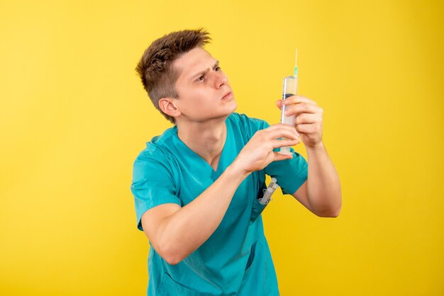 Junger männlicher Arzt der Vorderansicht im medizinischen Anzug, der Injektion auf gelbem Hintergrund hält