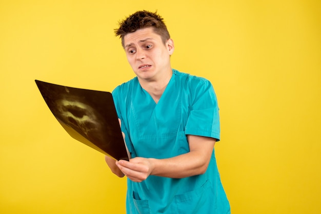 Junger männlicher Arzt der Vorderansicht im medizinischen Anzug, der Röntgenstrahl auf gelbem Hintergrund hält