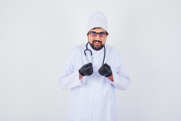 Junger männlicher Arzt in weißer Uniform posiert im Stehen und sieht fröhlich aus, Vorderansicht.