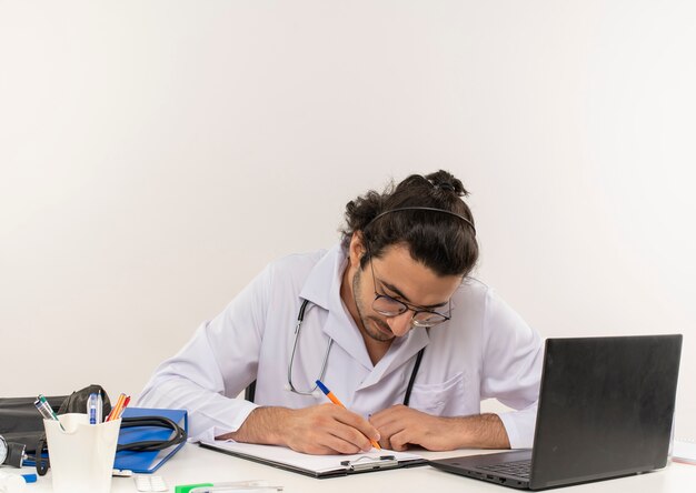 Junger männlicher Arzt mit medizinischer Brille, die medizinische Robe mit Stethoskop sitzend trägt