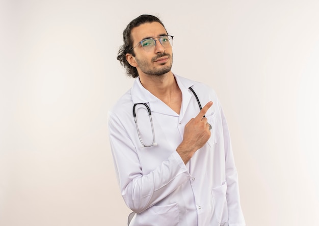 Junger männlicher arzt mit optischer brille im weißen gewand mit stethoskop zeigt zur seite