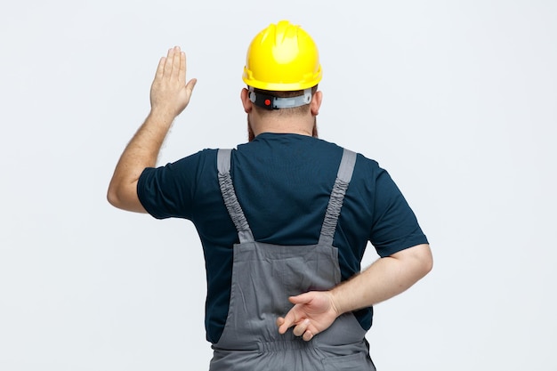 Junger männlicher Bauarbeiter mit Schutzhelm und Uniform, der hinter der Ansicht steht und Stoppgeste zeigt, während er die Finger hinter seinem Rücken kreuzt, isoliert auf weißem Hintergrund